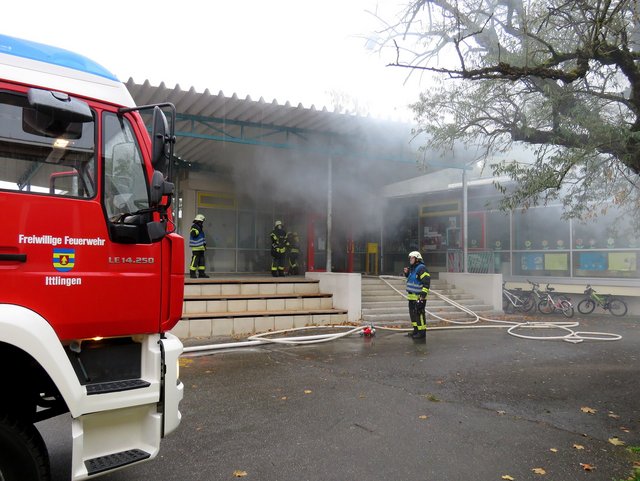 Feuerwehrfahrzeug vor Schulhaus und Feuerwehrmänner mit Schläuchen